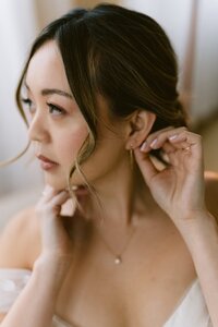 A bride in a white dress sitting by a window draped with sheer curtains.