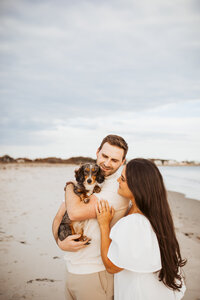 A couple hugging with their pet dog in their arms looking at their dog with a smile.