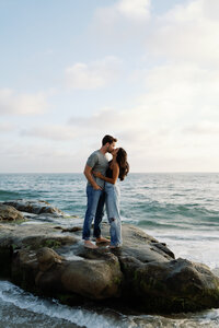 Couple sharing a kiss while standing on a large rock at Windansea Beach in San Diego, with waves lapping behind them and the afternoon light showing off the blue ocean creating a dreamy atmosphere.