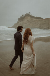 A couple elopes at Cape Kiwanda beach on a cold, rainy day. You couldn't wipe the smiles off their faces if you tried. They hold hands as they walk along the waters edge,  barefoot. She's holding the bottom of her muddied dress in one hand as she holds her grooms hand with the other. They resemble a real life Ariel and Prince Eric from the little mermaid.