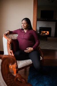 Mother to be sitting in a chair in a relaxed position with her hand resting on her belly, gazing out the window with a fire going in the background for pregnancy photos at home