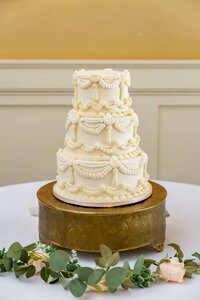 A three-tiered white wedding cake with ornate icing decorations sits on a gold cake stand, placed on a white table with green eucalyptus leaves and a pale pink rose.