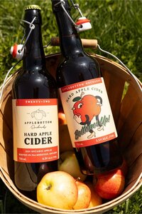 Two bottles of Applebottom Orchards hard apple cider on fresh apples in a wooden basket on the grass.