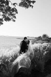 Couple shares a kiss during their wedding portraits, with the bride's veil blowing in the wind among the high grasses along the East Coast shoreline.