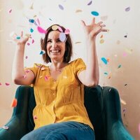 Gina Cooperman joyfully throwing confetti in the air while seated on a teal couch.