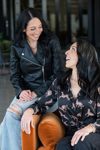 Two women laughing on an orange couch, engaged in a lively conversation about sales performance and techniques.