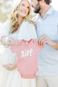 mom and dad holding baby girl pink onesie in gender reveal maternity photos at Cutter Creek Golf Course near Greenville, NC