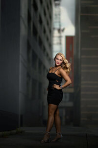A high school grad in a black dress stands in a windy alley