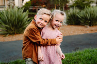 A close-up shot of a young boy and girl standing on a path in a park. The boy, wearing a brown shirt, is giving his sister, who is dressed in a pink dress, a sweet hug. Both children have bright smiles, and the background is filled with greenery.