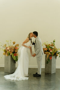 A bride and groom kiss on their wedding day after getting married inside a simple venue. Behind them are 2 big flower arrangements to each side of them.