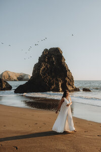 A woman in a flowing white dress walks along a sandy beach with a large, rocky outcrop in the background and birds flying overhead under a clear sky during a maternity photoshoot.