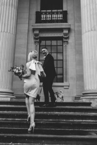 BRIDE AND GROOM STAND ON THE STAIRS OF THE OLD MARYLEBONE TOWNHALL WITH THEIR DOGS IN THEIR ARMS AS THEY GO DOWN THE STAIRS DURING CONFETTI