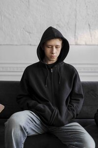 A masucline presenting teenager sits on a black couch, wearing a black zip-up hoodie pulled up. Their hands are tucked into both hoodie pockets, and they look downward toward their lap, with a sad expression.