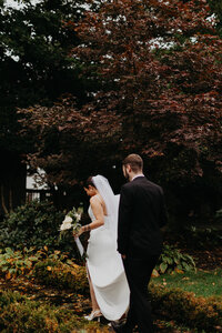 Bride strolls along an enchanting path, complemented by the groom gracefully holding her dress from behind – a captivating moment captured
