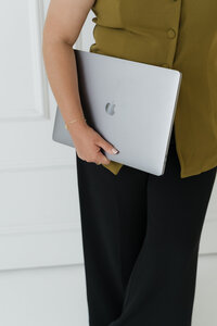 lose-up of woman in olive green top and black pants holding an Apple laptop against a white wall.