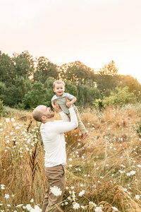 light-skinned dad holding up his toddler boy in the air in a field of golden grasses. Portland family photography session.