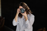 A woman with brown hair using a DSLR camera to take a photo. She wears a white shirt and stands in a room with a dark background, focusing on building her family photography business.