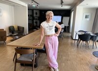 Tarzan wears pink baggy pants and a white cropped top, leaning against a long table in a very cool conference room, looking confident