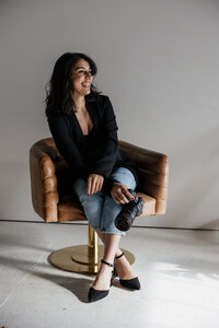 A woman with shoulder-length dark hair sits cross-legged on a brown swivel chair, smiling. A New England wedding photographer in her element, she wears a black top, light blue jeans, and black shoes, holding a camera in her lap. The scene is minimalistic with a plain background.