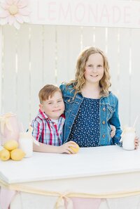Kids standing by a lemonade stand and holding a lemon and cup for a summer family photo session in North East, PA.