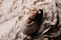 A photo of a newborn girl who is swaddled in the potato sack style. The wrap and the backdrop are both textured and khaki colored. The baby is wearing a khaki colored bow on her head. The image is shot from the dark side, showcasing bright highlights and dark shadows to enhance all of the details of the newborn. Her gorgeous dark skin is glowing as she sleeps peacefully. Photographed by Mobile, AL Newborn Photographer