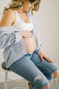 Pregnant mom sits on stool in studio and looks at bare belly