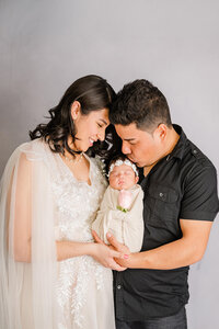 Parents in formal attire, cuddle close as they hold a newborn baby wrapped in a beige fabric between them.