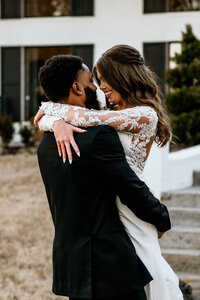 The groom is lifting his bride into the air as she wraps her arms around his shoulders, bringing their faces closer together