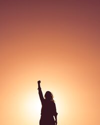 The silhouette of a woman with her fist raised stands against a sky made orange by the sunset.