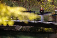 High school senior in black SENIORS hoodie standing on bridge looking into the distance, framed by fall trees and golden light