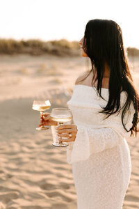bride holding wine glasses on beach during sunset beach engagement session