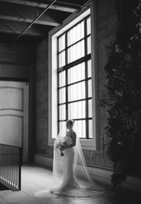 Black and white photo of a bride standing by a window at The Switch House in Philadelphia, bathed in soft natural light with a contemplative expression.