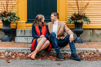 Couple in peacoats sit on the sidewalk in front of a historic home