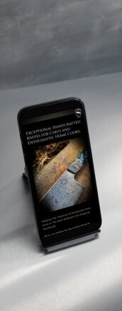 A smartphone displaying a mobile-optimised webpage for Cuttlebrook Knives. The screen shows an image of an intricately designed chef knife with text that reads, "Exceptional handcrafted knives for chefs and enthusiastic home cooks." The phone is placed on a sleek, metallic surface, with a blurred background.