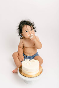 One Year old baby milestone photo session with cake. Baby is in a blue diaper cover and nothing on the top. He has a beautiful, white cake in front of him and is putting some cake in his mouth and has frosting all over his hand. He is looking at the camera with a very serious look on his face. He has longer dark curly hair.