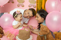A joyful family celebrates a baby's birthday, captured by a Jacksonville cake smash photographer. The baby, in a pink tutu and floral headband, smiles at the themed cake. The parents, surrounded by pink balloons and teddy bears, express happiness amid pastel pink and brown decor that sets a festive tone.