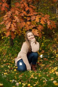 A young pre-teen girl smiling and crouching down with the bright orange fall colors behind her.