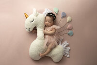 Newborn baby rides on the back of a stuffed unicorn holding onto balloons.