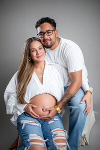 Young hispanic couple smiling and holding pregnant belly for a maternity session in the studio. 