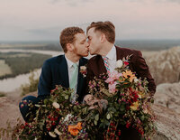A gay couple in wedding attire sit on the cliffside of Stouts Point at Petit Jean State Park. They are leaned into each other for a kiss while they hold a very large bouquet draped over both their laps.