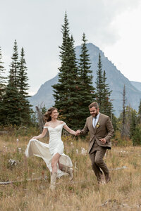 Bride and groom run hand in hand through a mountain meadow at Glacier National Park, surrounded by pine trees and golden light.