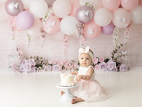 girl in pink dress sitting with cake for first birthday cake smash