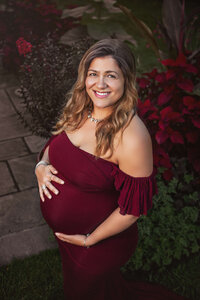 pregnant mother weaing a burgundy maternity gown holding her baby bump in a flower garden