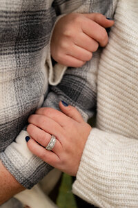 Two people stand closely, adorned in cozy plaid and knitted sweaters. A large, sparkling ring catches the eye on one's hand as they embrace warmly. Captured with the finesse of a New England photographer, this moment exudes fine art elegance and documentary-style intimacy.