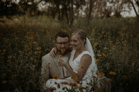 Bride is sitting on the groom's lap in a field surrounded by yellow flowers.