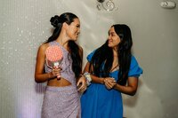 Two young women smile and link arms during a celebration at SoHo63, a Chandler wedding and event venue. One guest holds a light-up prop paddle while wearing a sparkly lavender dress, highlighting the playful and vibrant energy of events hosted at SoHo63.