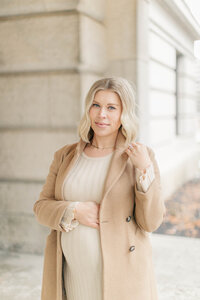 Expecting mother stands beside a stone column wearing a cream dress and tan coat during maternity session at The Nelson in Kansas City