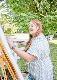 Image of wedding artist Elyse Stokes laughing while painting on canvas and easel while doing a live wedding painting