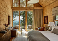 A cozy bedroom with wooden walls and ceiling, featuring a large bed, a dresser, and a seating area. Large windows and sliding glass doors lead to a balcony with outdoor furniture, overlooking a forested landscape in the background.