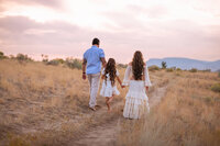 A family of three hold hands while walking away from the camera. A beautiful sunset is in front of them in Valleyview Nature Park in Kamloops.